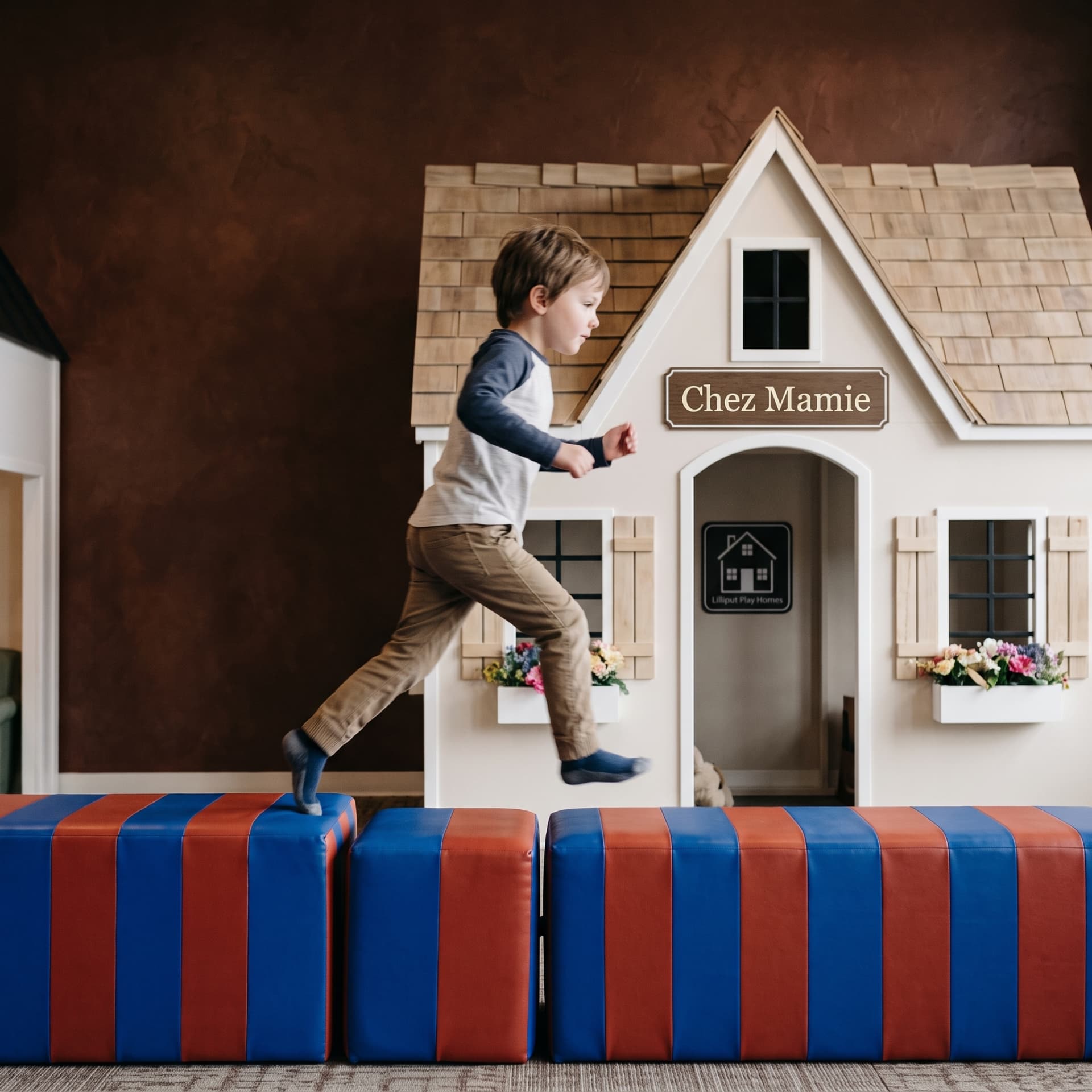 Child leaping across red and blue striped play blocks in front of a Chez Mamie playhouse